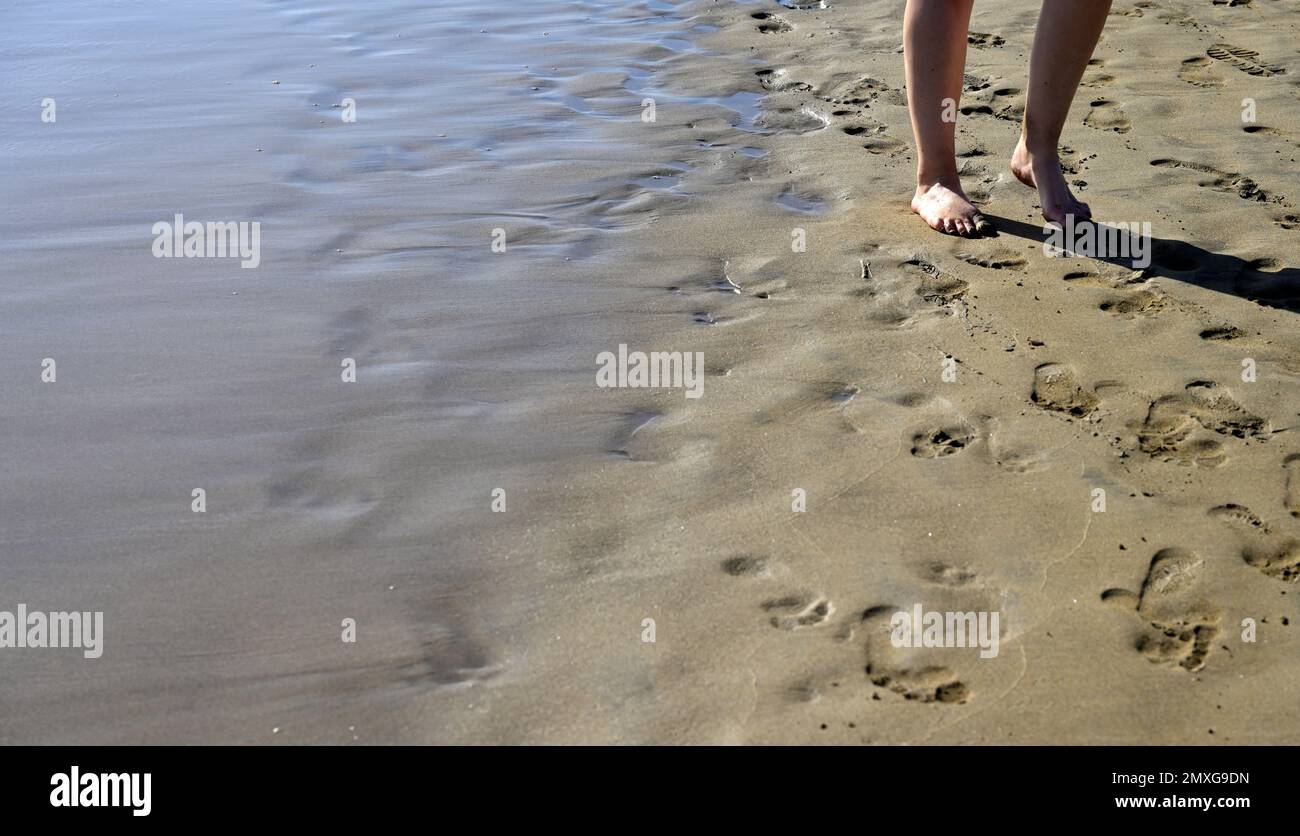 Gambe e piedi di una persona a piedi nudi che cammina lungo la spiaggia sabbiosa al bordo delle acque Foto Stock