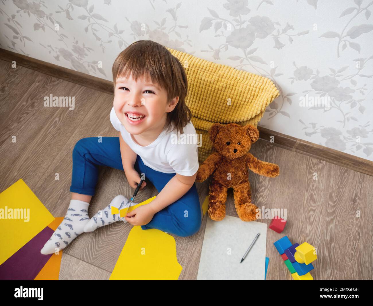 Il bambino impara a tagliare carta colorata con le forbici. Il bambino si siede sul pavimento nella stanza dei bambini con i blocchi del giocattolo e l'orso del teddy. Corsi educativi per bambini Foto Stock