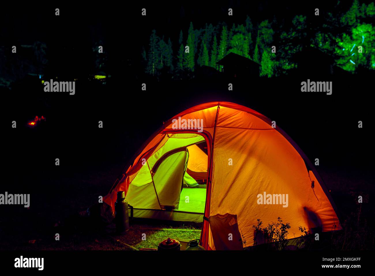 Una tenda turistica con un ingresso aperto si illumina sullo sfondo di sagome di persone intorno al fuoco e case. Foto Stock