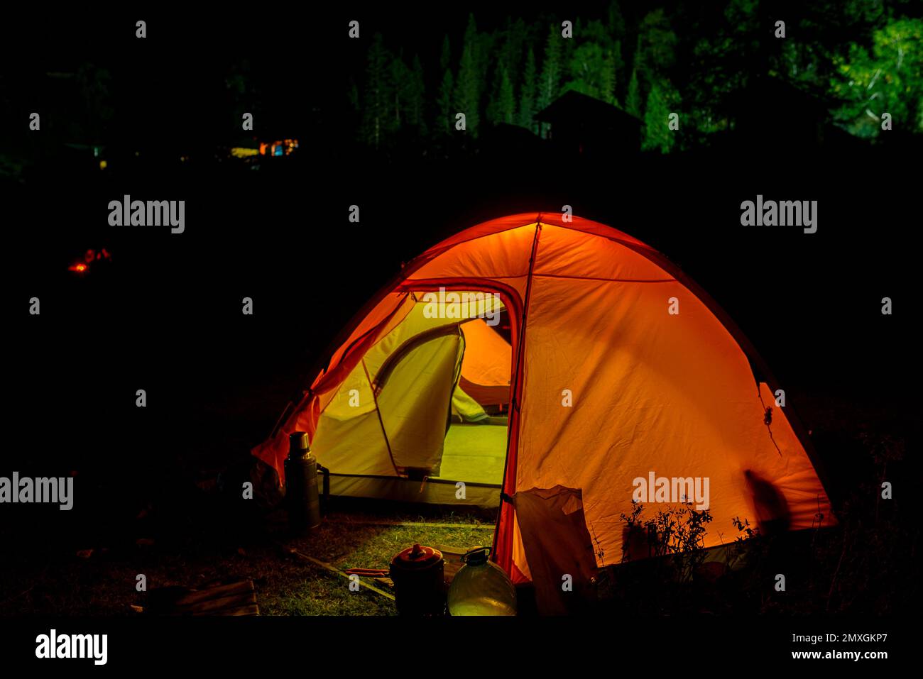 Una tenda turistica con un ingresso aperto si illumina sullo sfondo di sagome di persone intorno ad un fuoco e case con luce verde. Foto Stock