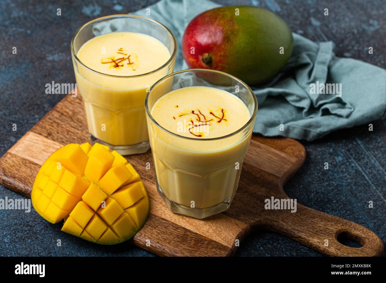 Sani Ayurveda indiano bere mango lassi in due bicchieri su tavolo rustico in cemento con mango fresco tagliato maturo, bevanda mista gialla fatta di mango fru Foto Stock