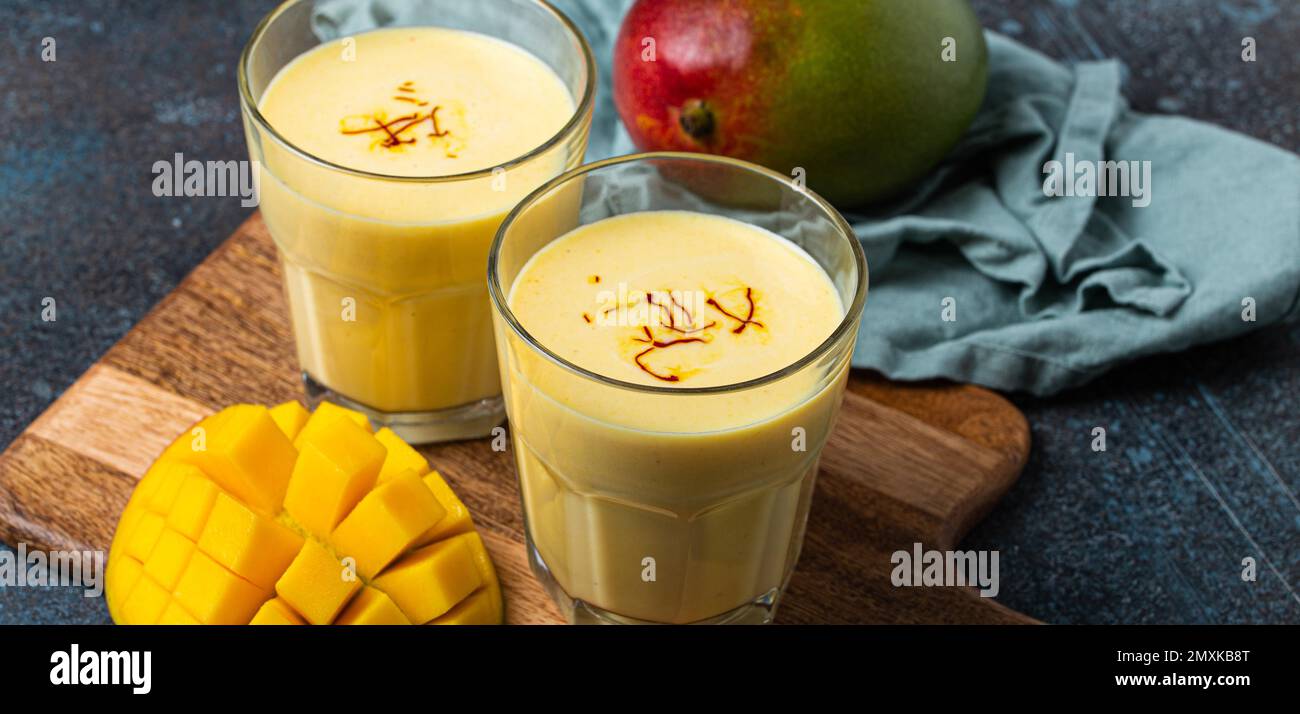 Sani Ayurveda indiano bere mango lassi in due bicchieri su tavolo rustico in cemento con mango fresco tagliato maturo, bevanda mista gialla fatta di mango fru Foto Stock