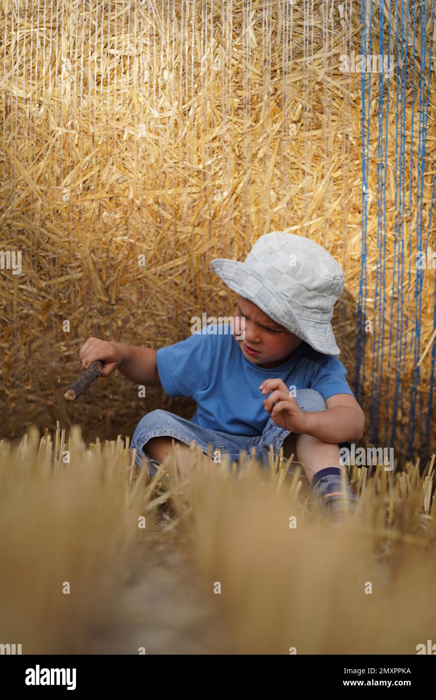 Piccolo adorabile bambino con grandi occhi marroni in un cappello siede in  un campo vicino a fienate al tramonto in estate, e guarda un filo d'erba.  Selettivo Foto stock - Alamy