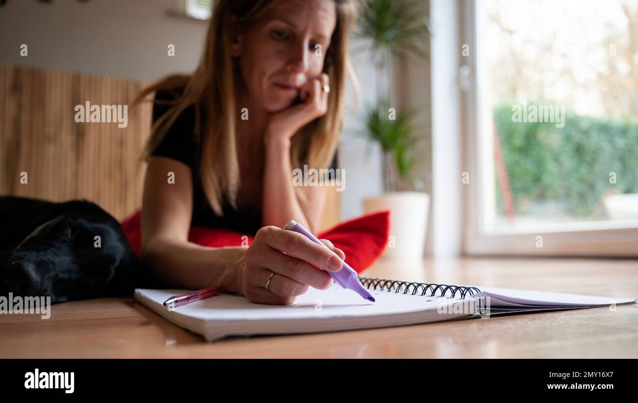 Giovane donna che legge e studia sdraiata sul pavimento di un soggiorno evidenziando informazioni importanti con un pennarello viola in un notebook. Con il suo cane lyin Foto Stock