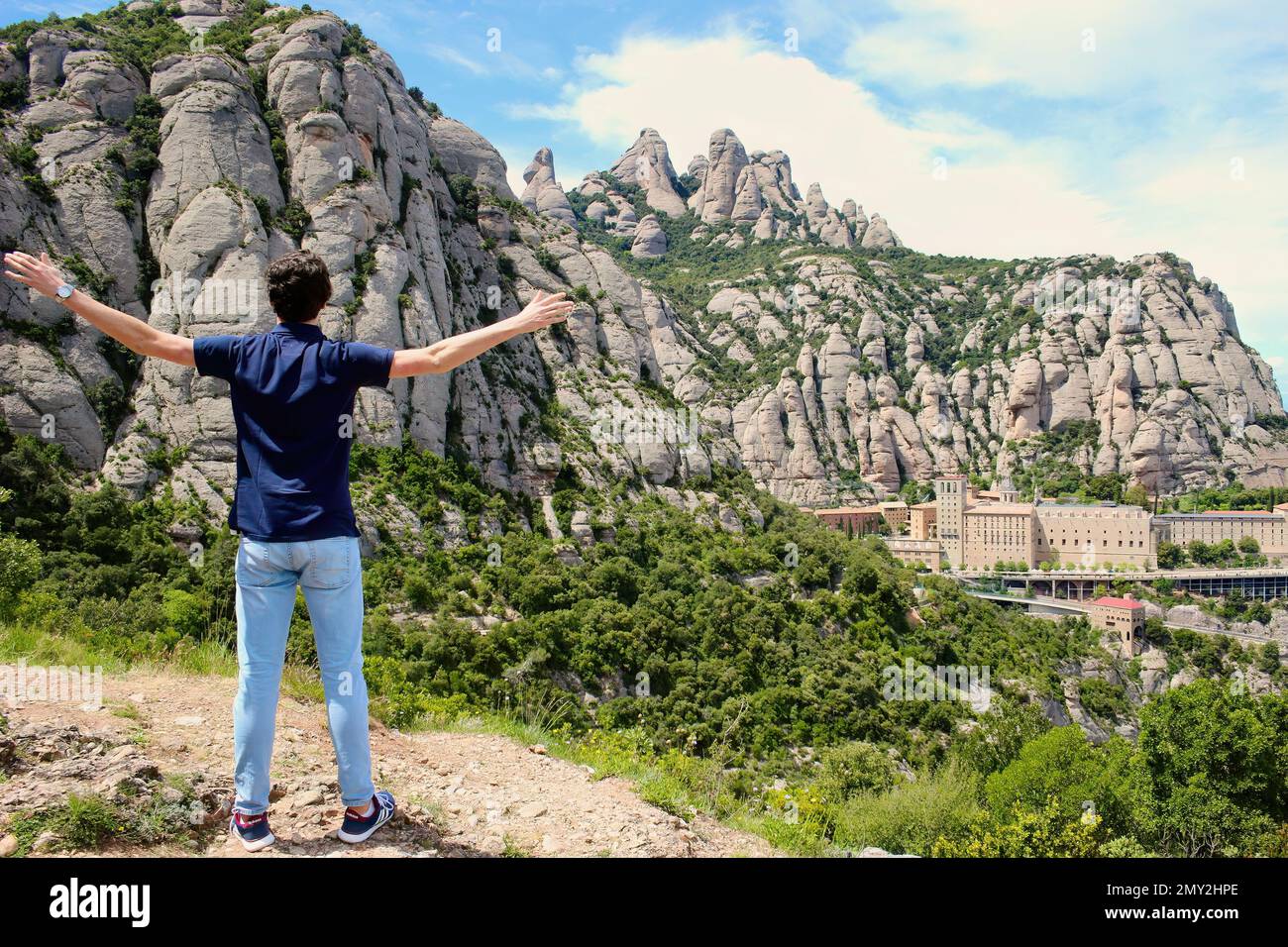 Vista del paesaggio con giovane uomo con ritorno alla macchina fotografica e le braccia estese Montserrat monastero benedettino Dawn con luce solare forte e ombre profonde Foto Stock