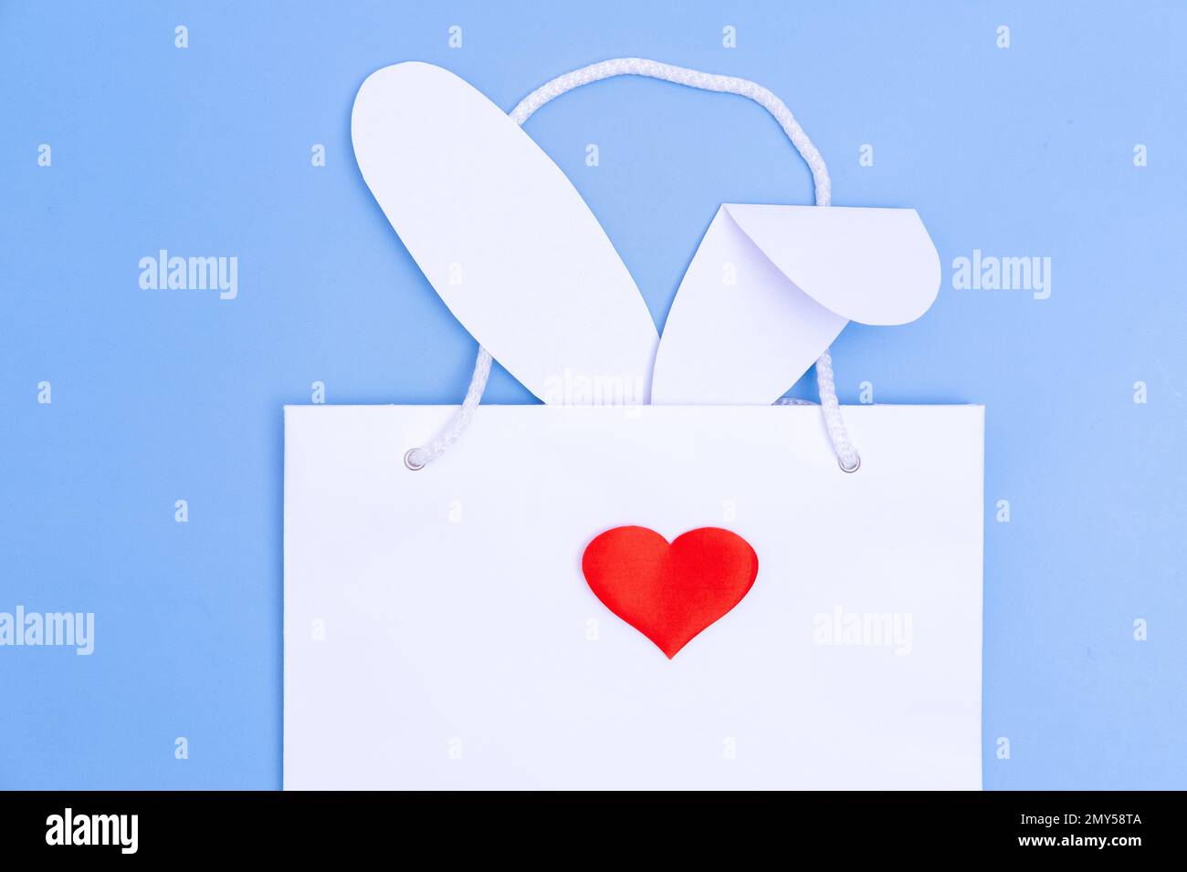 Shopping per Pasqua. Borsa della spesa in carta bianca con orecchie bianche e cuore rosso come naso su sfondo blu. Concetto regali di Pasqua, preparazione, Foto Stock