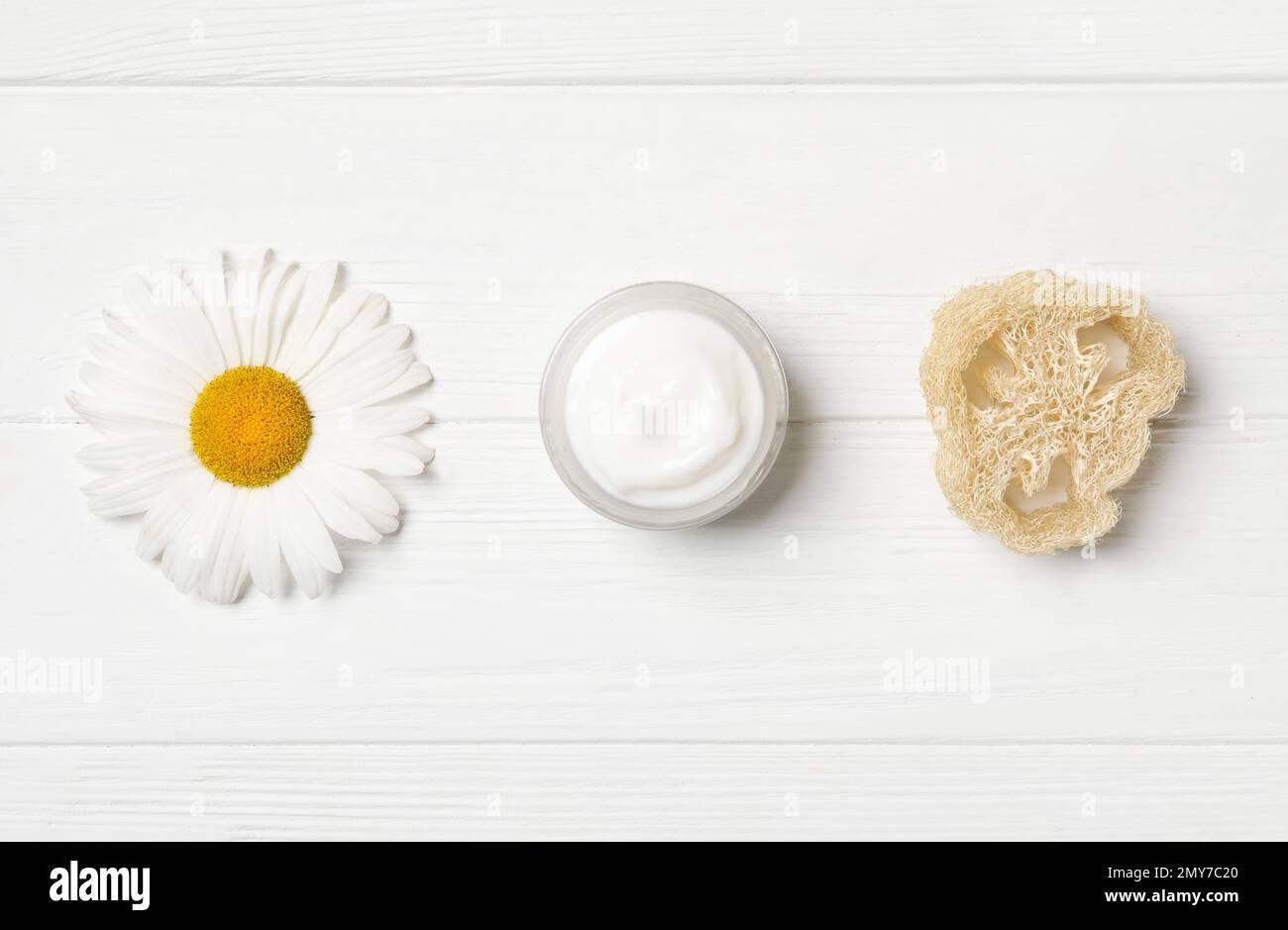 Vasetto di crema, loofah e camomilla su tavolo di legno bianco, piatto Foto Stock