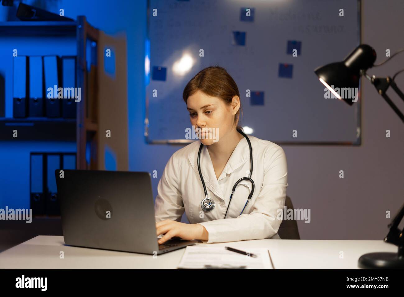 Attraente dottore in camice bianco seduto al tavolo con laptop durante il turno di notte. Spazio di copia Foto Stock
