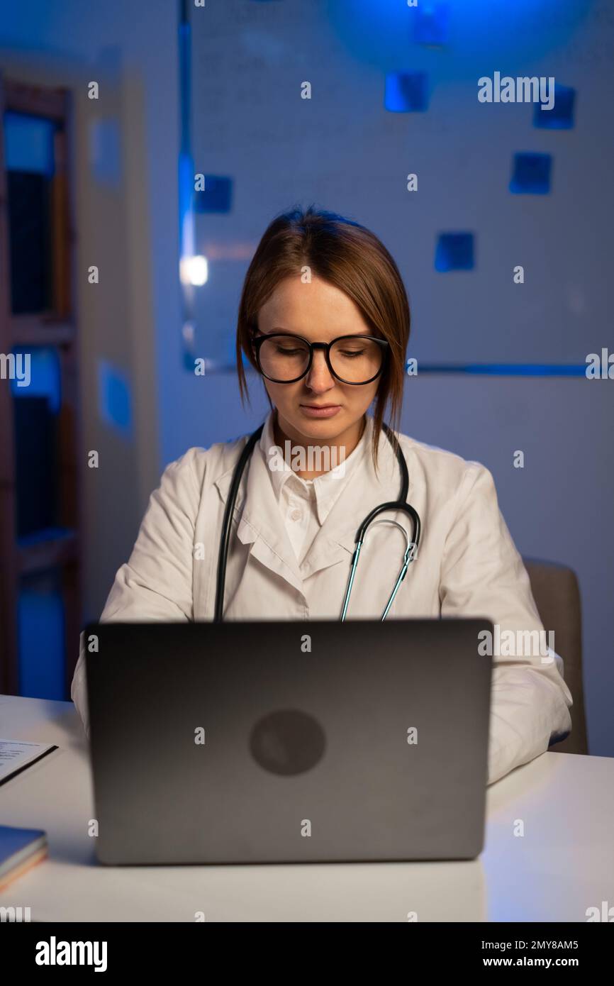 Dottore in camice bianco e occhiali che scrivono sul computer portatile in ospedale durante il turno di notte. Assistenza sanitaria, tecnologia e medicina Foto Stock