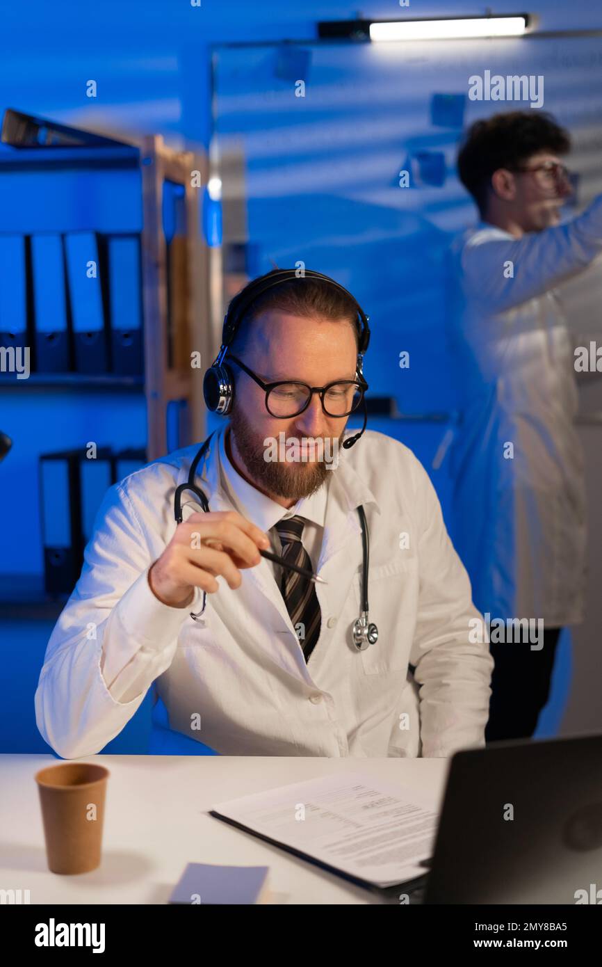 Medico con cuffie che consulta il paziente online alla scrivania in clinica durante il turno di notte, il suo collega giovane medico in piedi sullo sfondo. Foto Stock