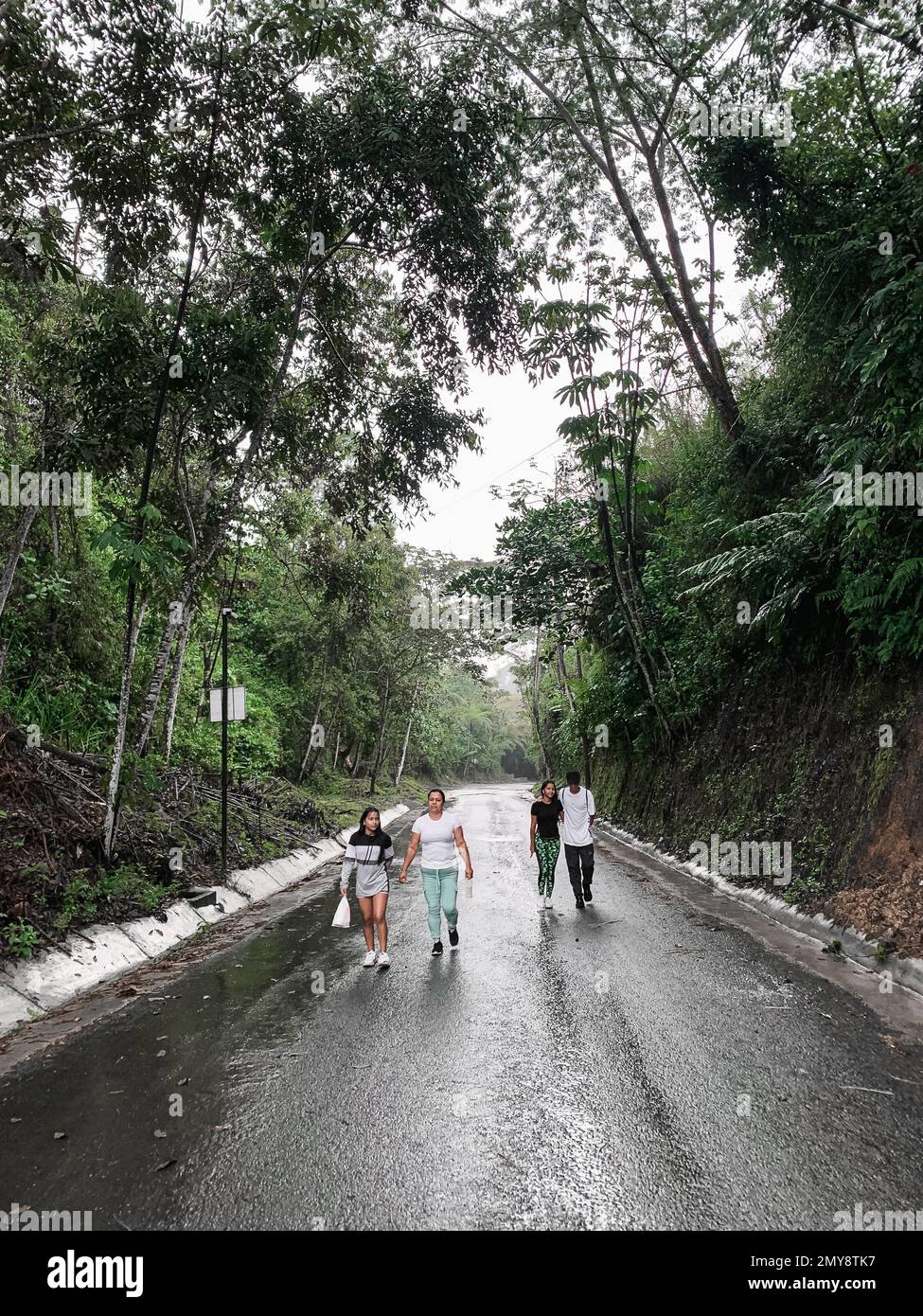 madre latina che cammina con i suoi figli su una strada solitaria in una giornata di pioggia, circondata dalla natura. Foto Stock