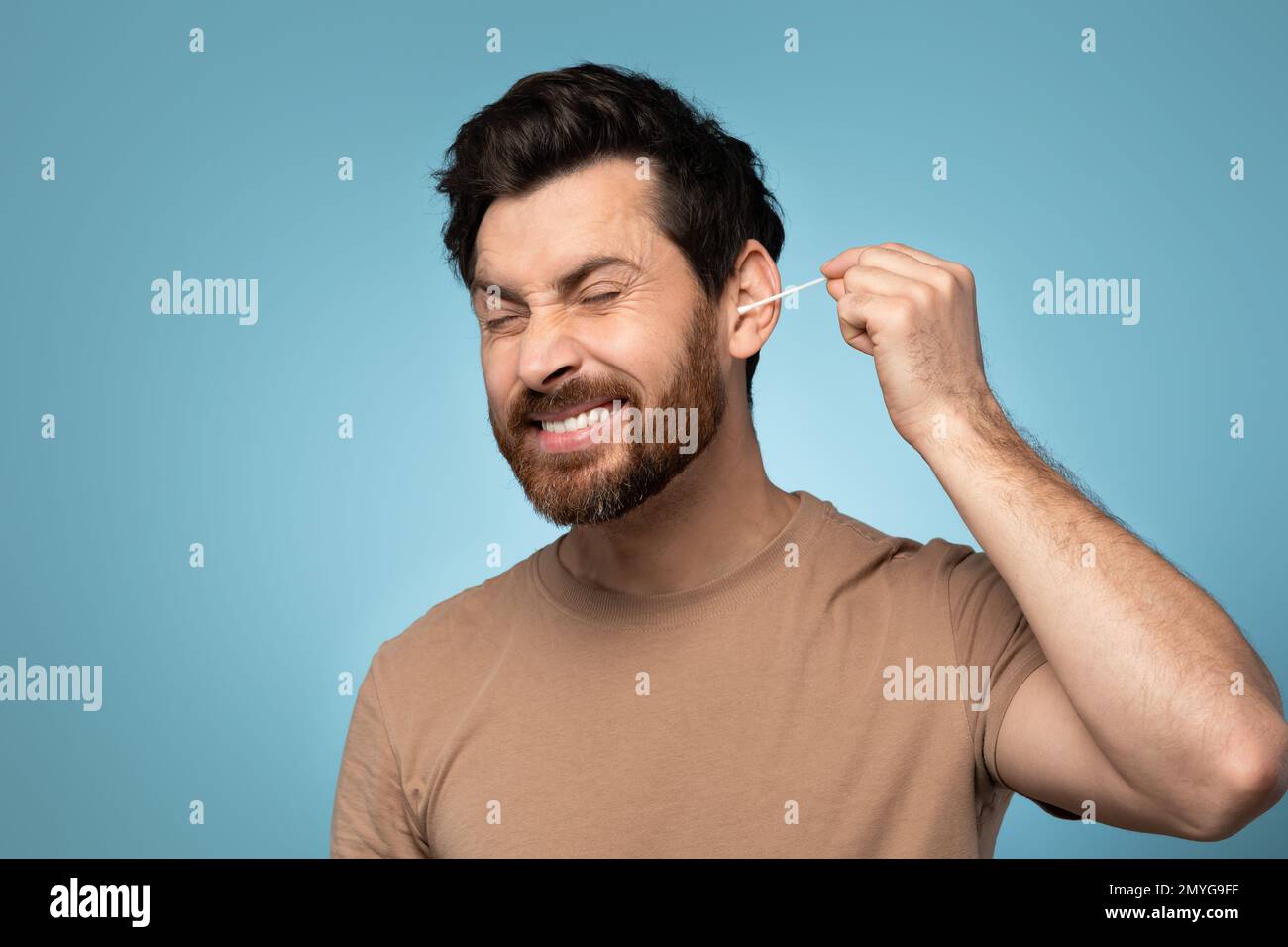 Routine di igiene quotidiana. Uomo bearded divertente pulire le orecchie con bastoncino di cotone, facendo mattina routine di bellezza, sfondo blu Foto Stock