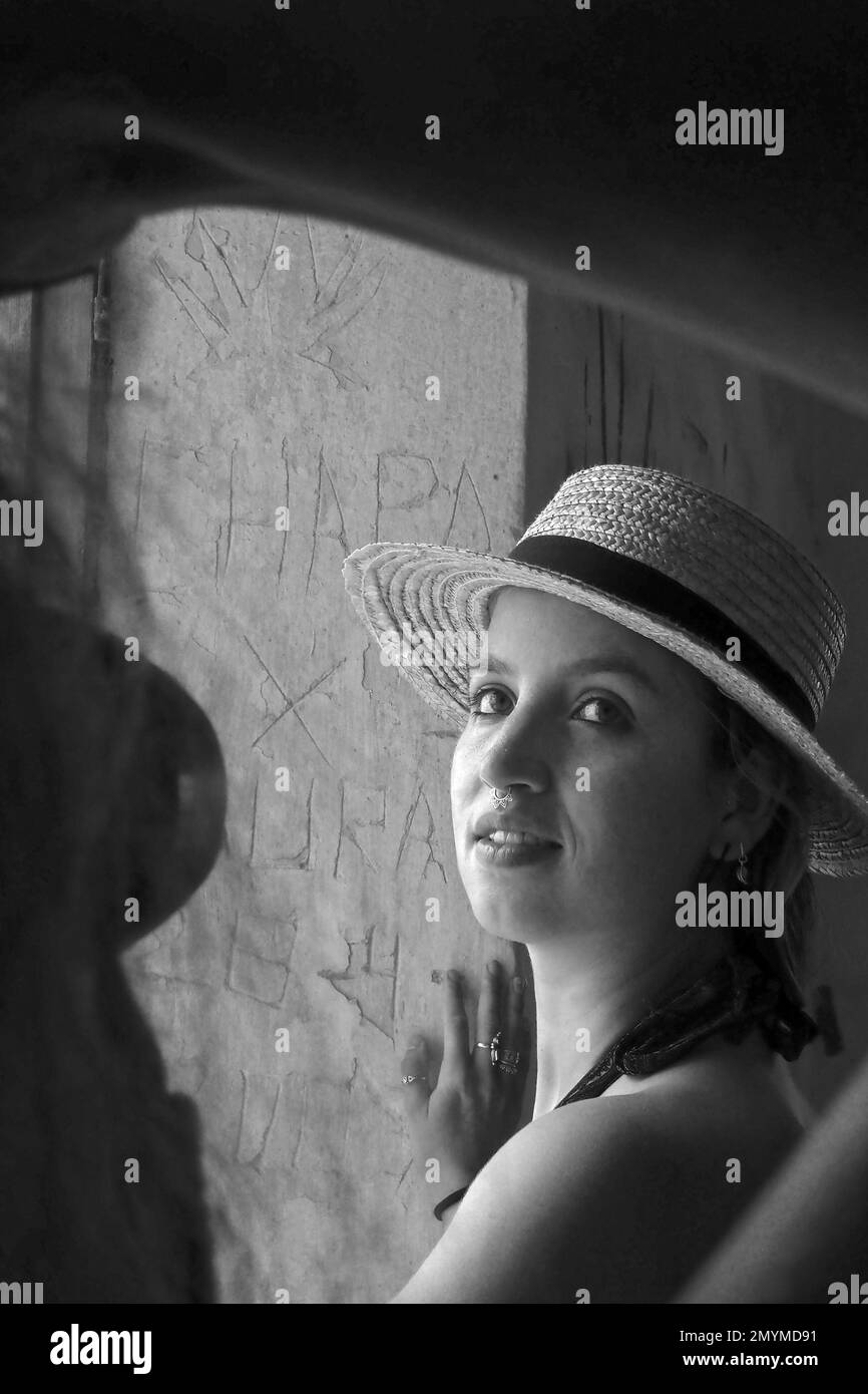 Giovane donna con cappello di paglia guarda curiosamente in macchina fotografica, foto in bianco e nero Foto Stock