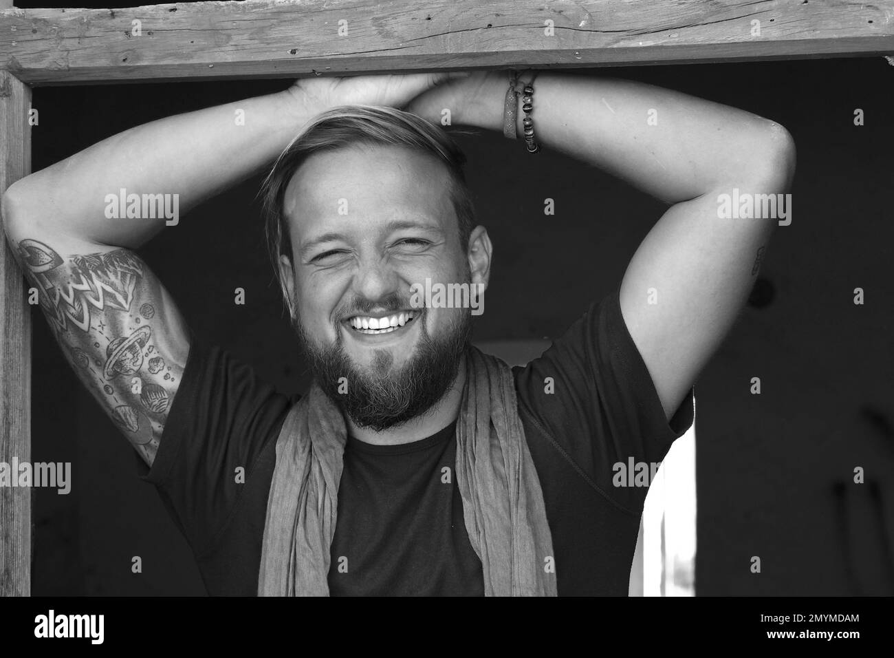 Giovane uomo con barba e tatuaggio sul braccio si trova nella porta e ride felicemente, foto in bianco e nero Foto Stock