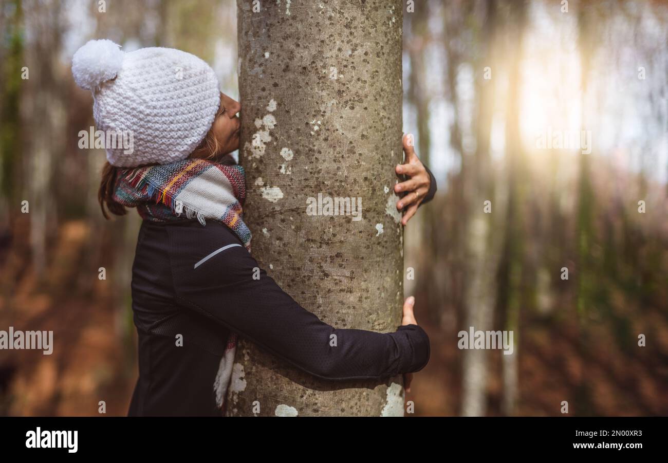 donna di mezza età in cappuccio bianco abbraccia e bacia un tronco d'albero al tramonto. concetto di connessione con la natura. balneoterapia. Foto Stock