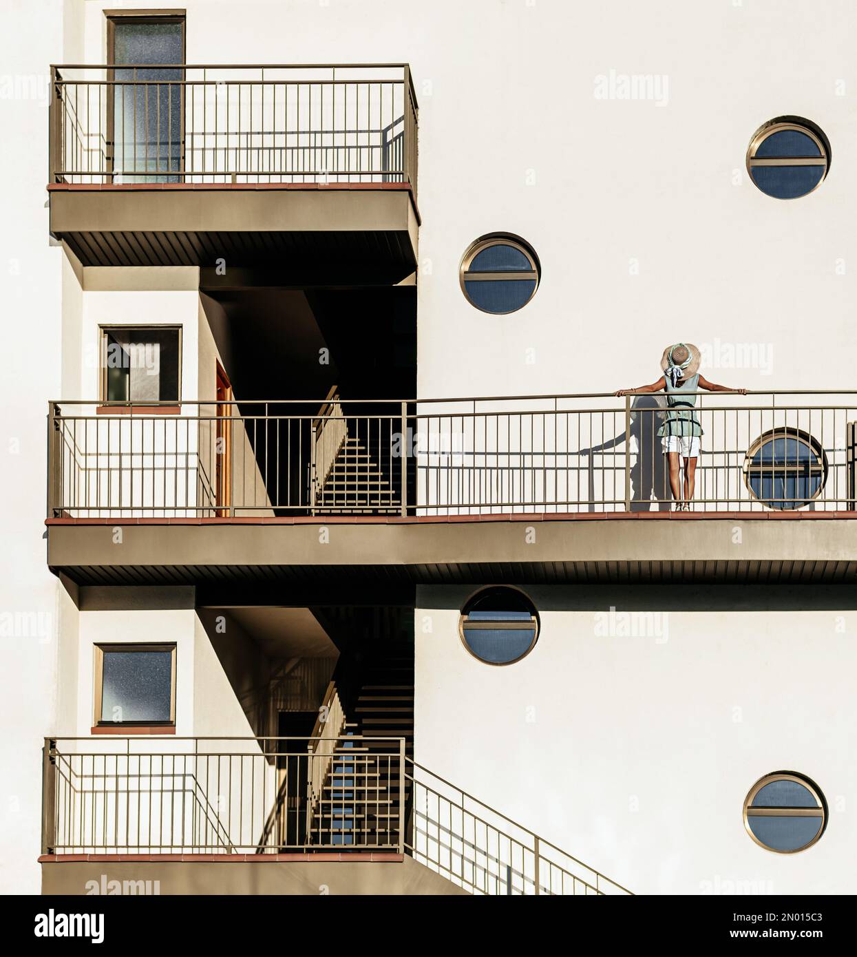 una donna di mezza età con un cappello sulla schiena si appoggia alla ringhiera di un edificio moderno con finestre rotonde. concetto di minimalismo e architettura urbana. Foto Stock