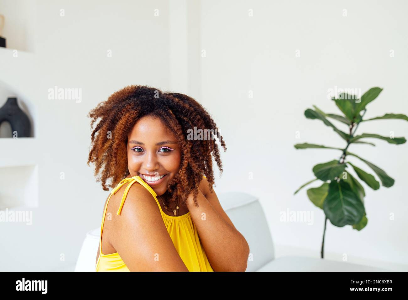 Bella curvy oversize giovane afto americano donna in un abito giallo seduto su un divano a casa. Corpo positivo elegante donna sorridente hanno tempo di relax. Foto Stock