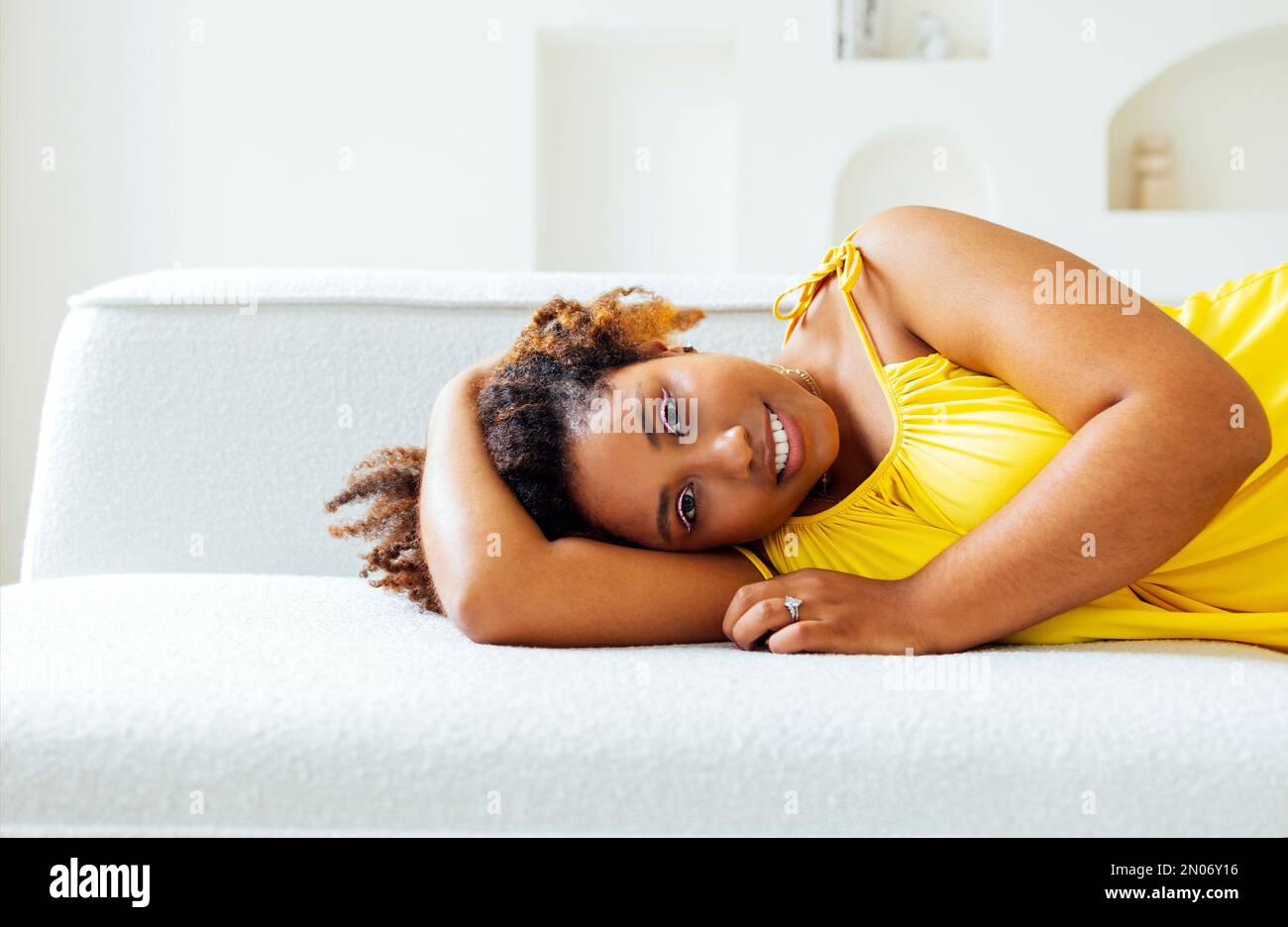 Bella curvy oversize giovane afto americano donna in un abito giallo si trova su un divano a casa. Corpo positivo elegante donna sorridente hanno tempo di relax. BOD Foto Stock