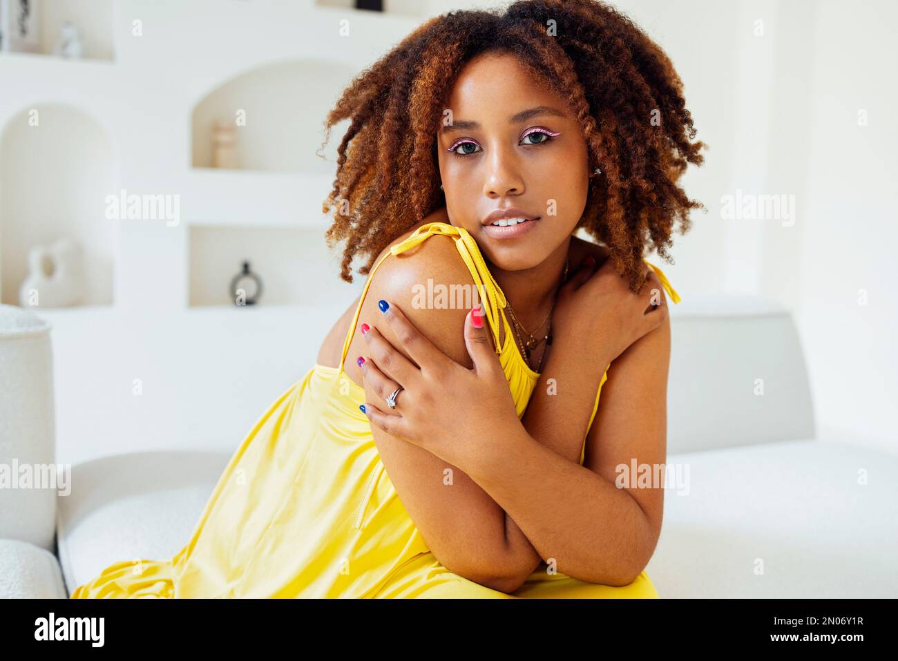 Bella curvy oversize giovane afto americano donna in un abito giallo seduto su un divano a casa. Corpo positivo elegante donna sorridente hanno tempo di relax. Foto Stock