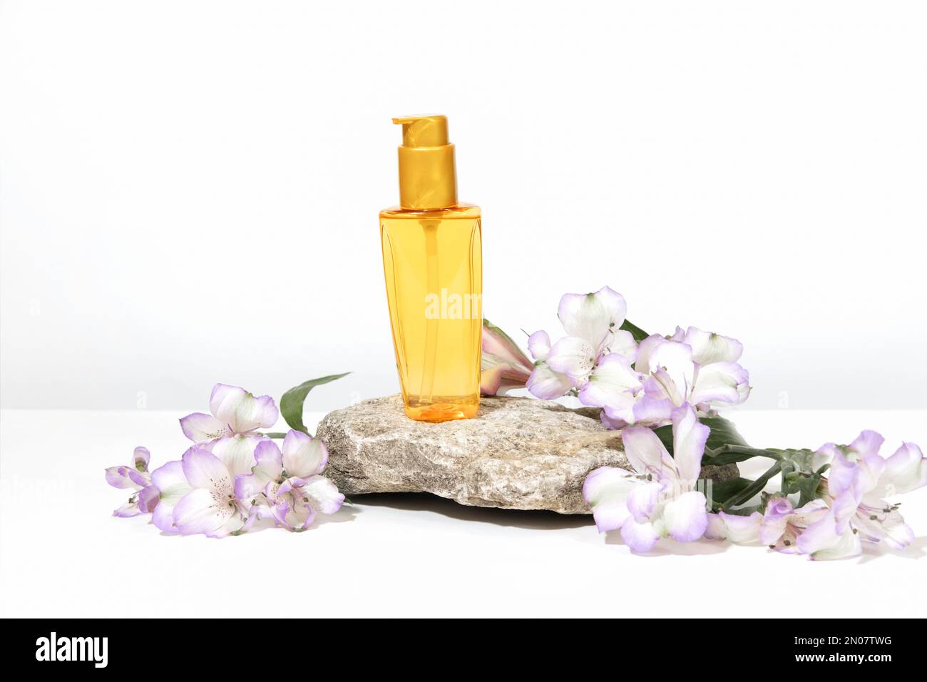 Una bottiglia trasparente di olio per la protezione solare, abbronzatura o cura dei capelli, su un piedistallo da podio. Sfondo grigio bianco con ombre dure, fiori e piante Foto Stock