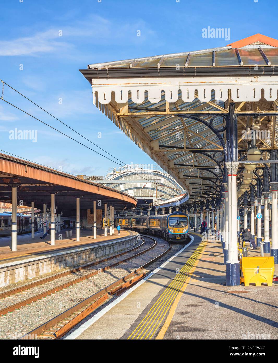 Vista di una stazione ferroviaria con rotaie che conducono verso un arco di ferro. Una piattaforma è coperta da una tettoia del 19th ° secolo e un treno si trova accanto a un plateau Foto Stock