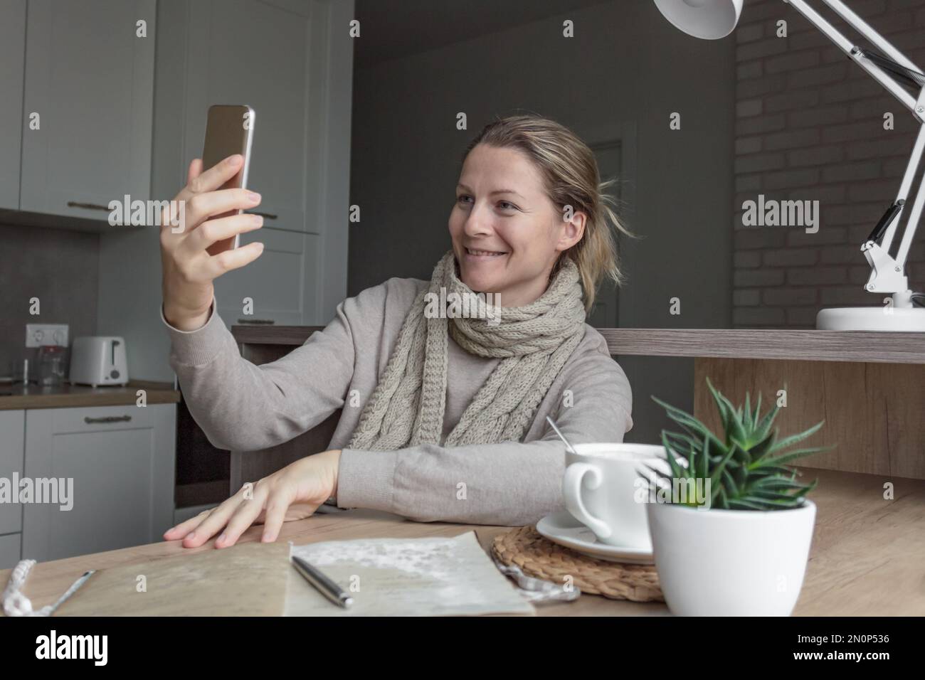 Una donna in un maglione e una sciarpa a maglia in cucina siede ad un tavolo di legno e parla su una videochiamata al telefono. Foto Stock