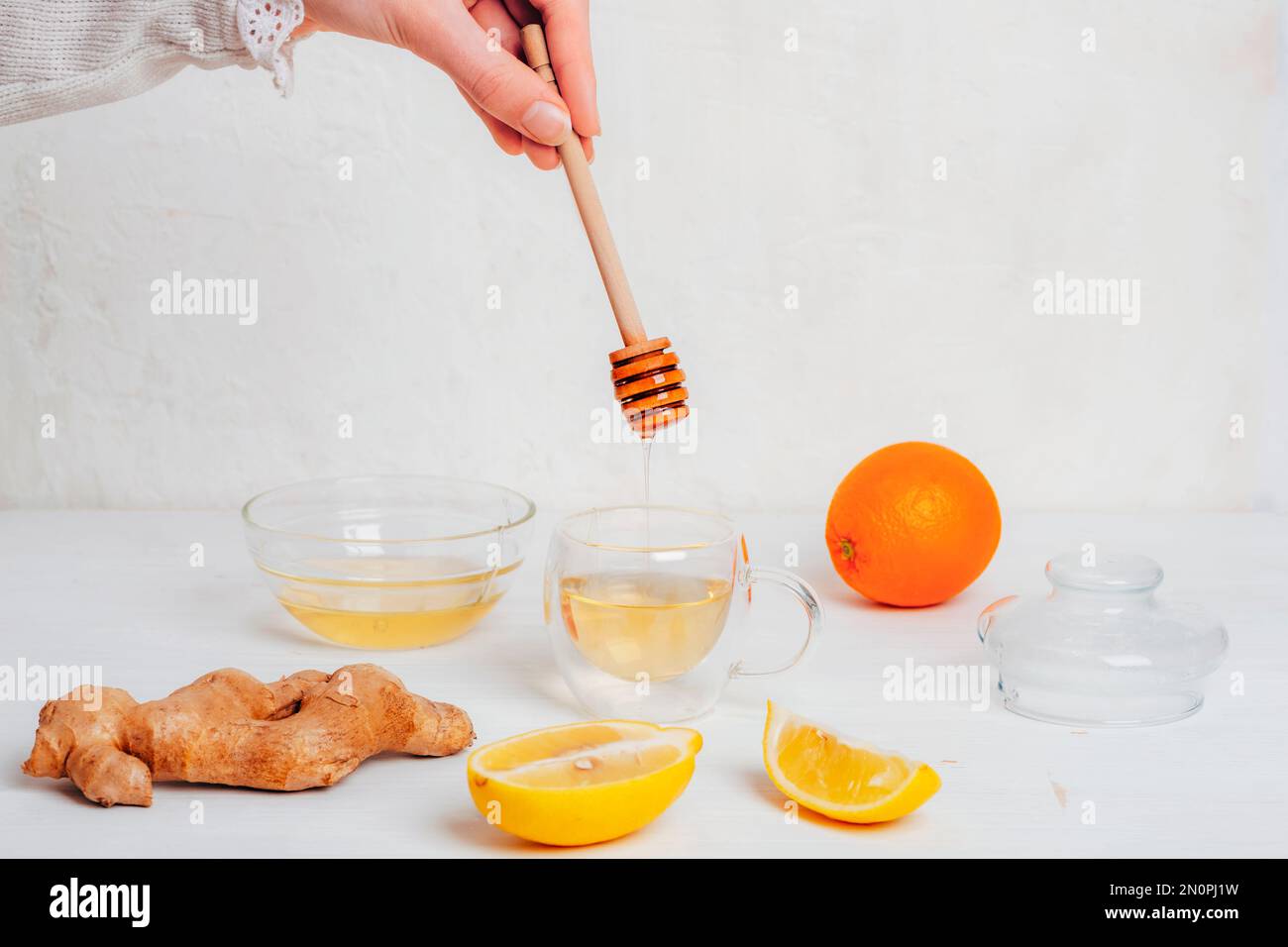 Bicchiere con tè all'arancia, al limone e allo zenzero e miele che si versa in una tazza. Ingredienti su tavola di legno bianco. Foto Stock