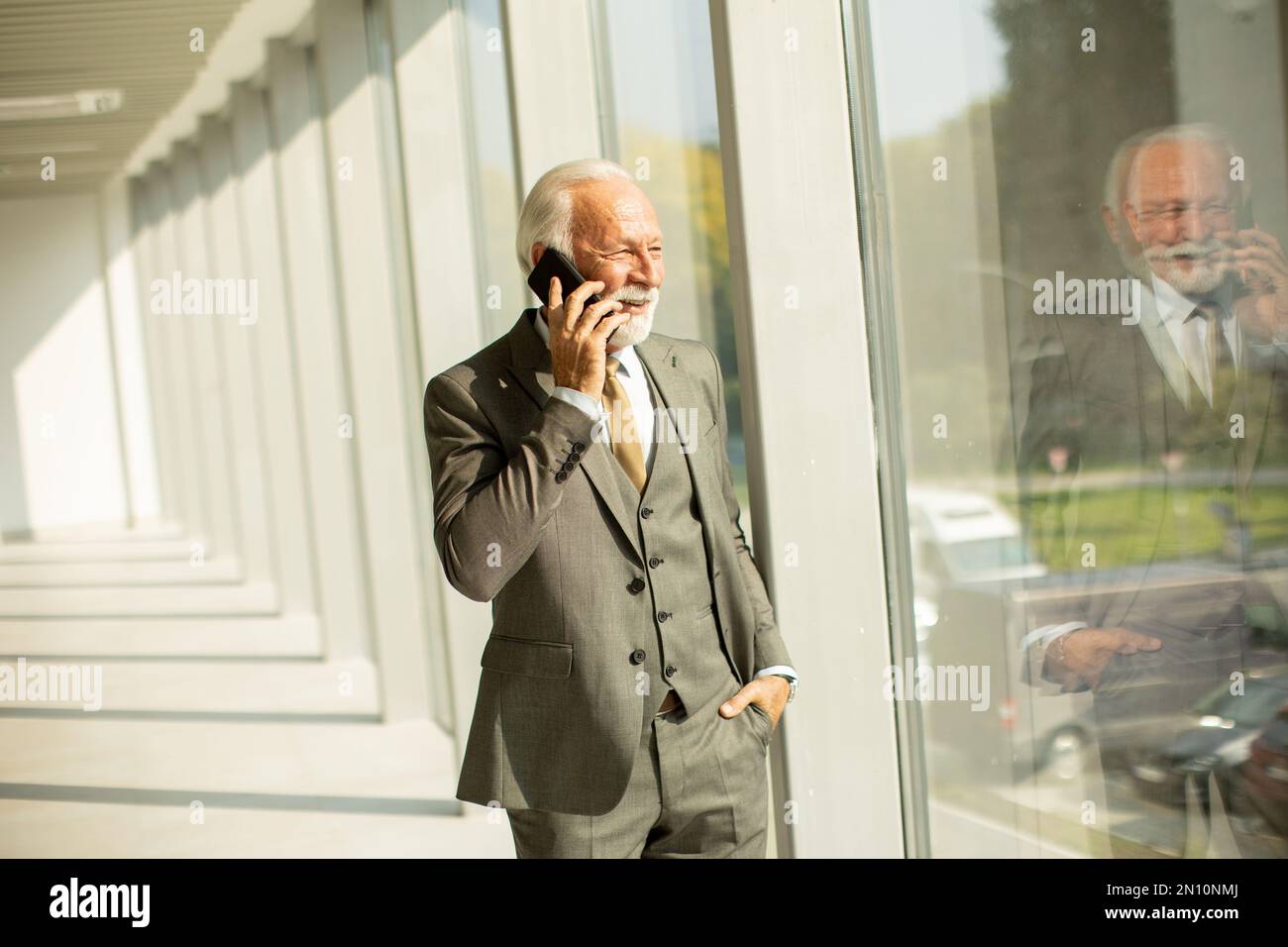 Un uomo d'affari senior si trova in un corridoio dell'ufficio, concentrato sul suo telefono cellulare. È vestito in abbigliamento formale, che trasuda fiducia e professionalità Foto Stock