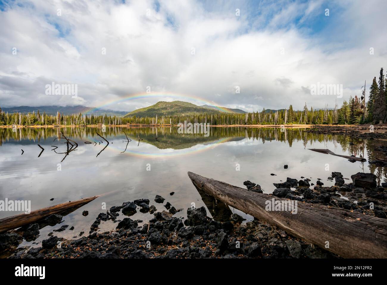 Tronchi di alberi che si aggettano in acqua, arcobaleno in nuvole scure sopra una foresta, riflesso nel lago, Sparks Lake, Oregon, Stati Uniti, Nord America Foto Stock