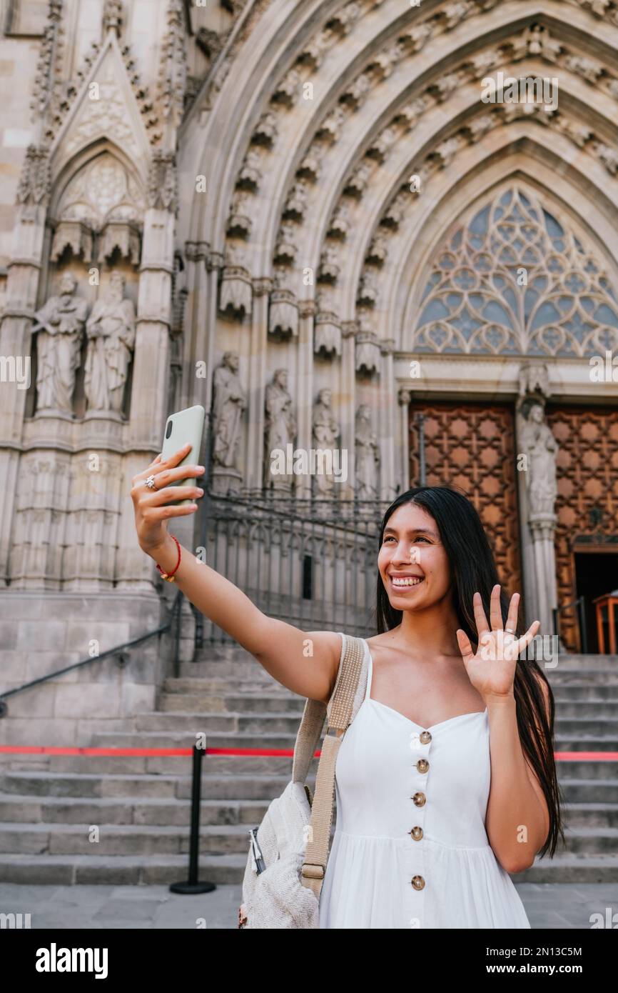 Allegra donna ispanica in abito bianco con zaino che agita la mano e sorride mentre si effettua una videochiamata via smartphone fuori dalla cattedrale medievale di Barcellona, Spagna Foto Stock