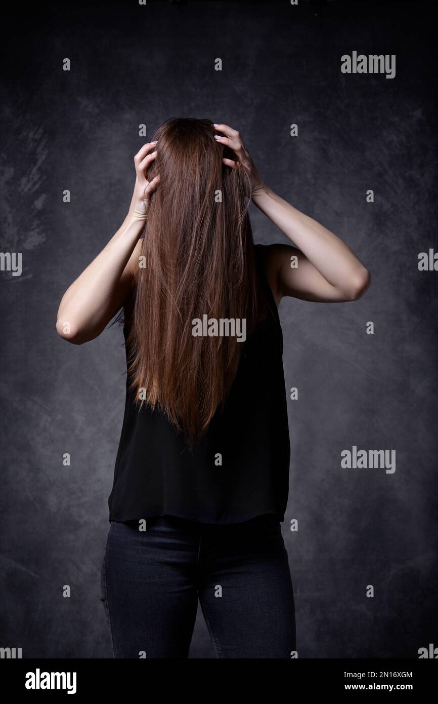 Giovane donna con capelli lunghi che tiene le mani sulla testa. I capelli coprono il viso. Sfondo grigio. Concetto di cefalea ed emicrania. Foto Stock
