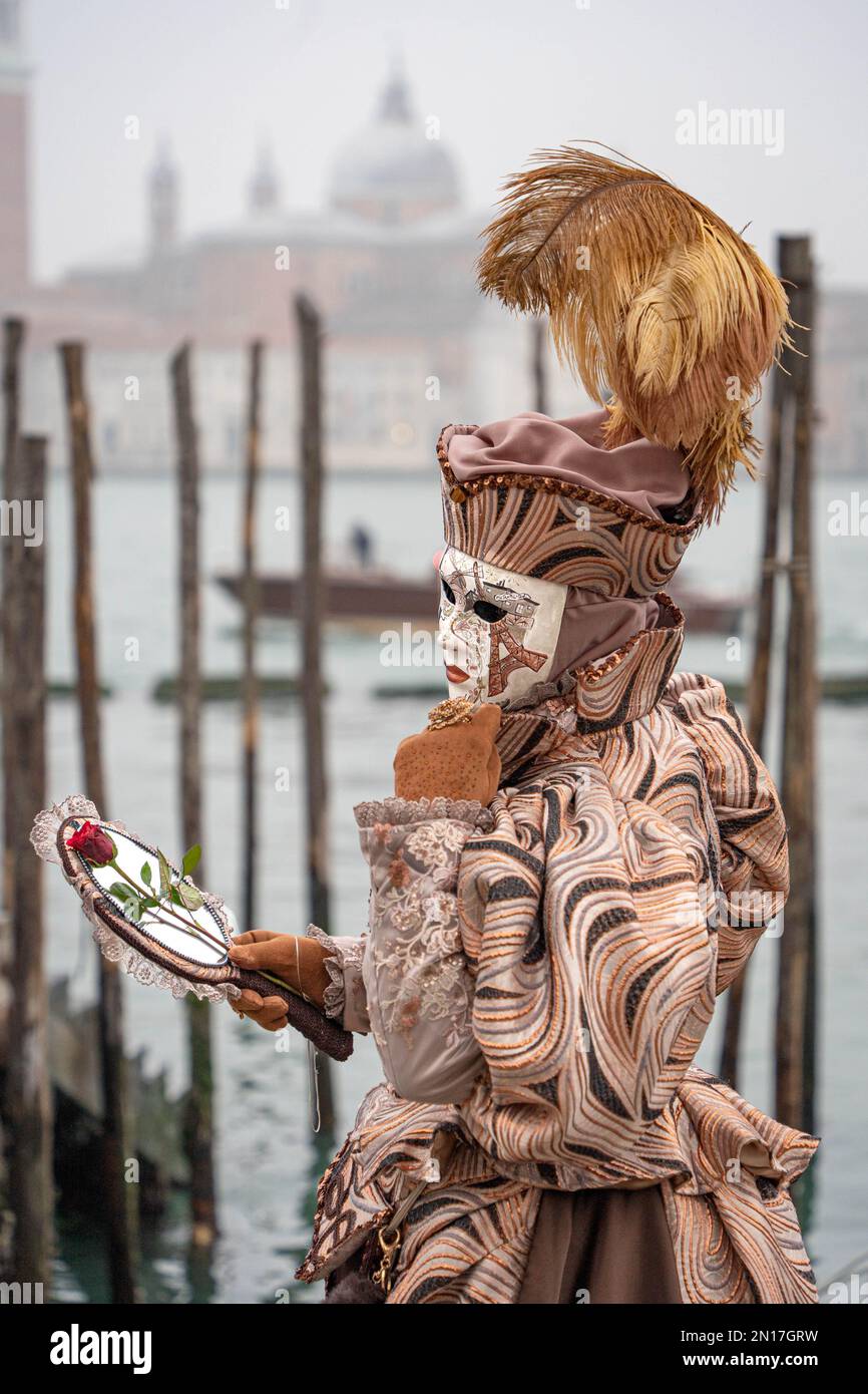 Una donna in costume da carnevale, una maschera, un cappello con piume, uno specchio e un fiore in mano, si trova sullo sfondo del molo e della Basilica Foto Stock