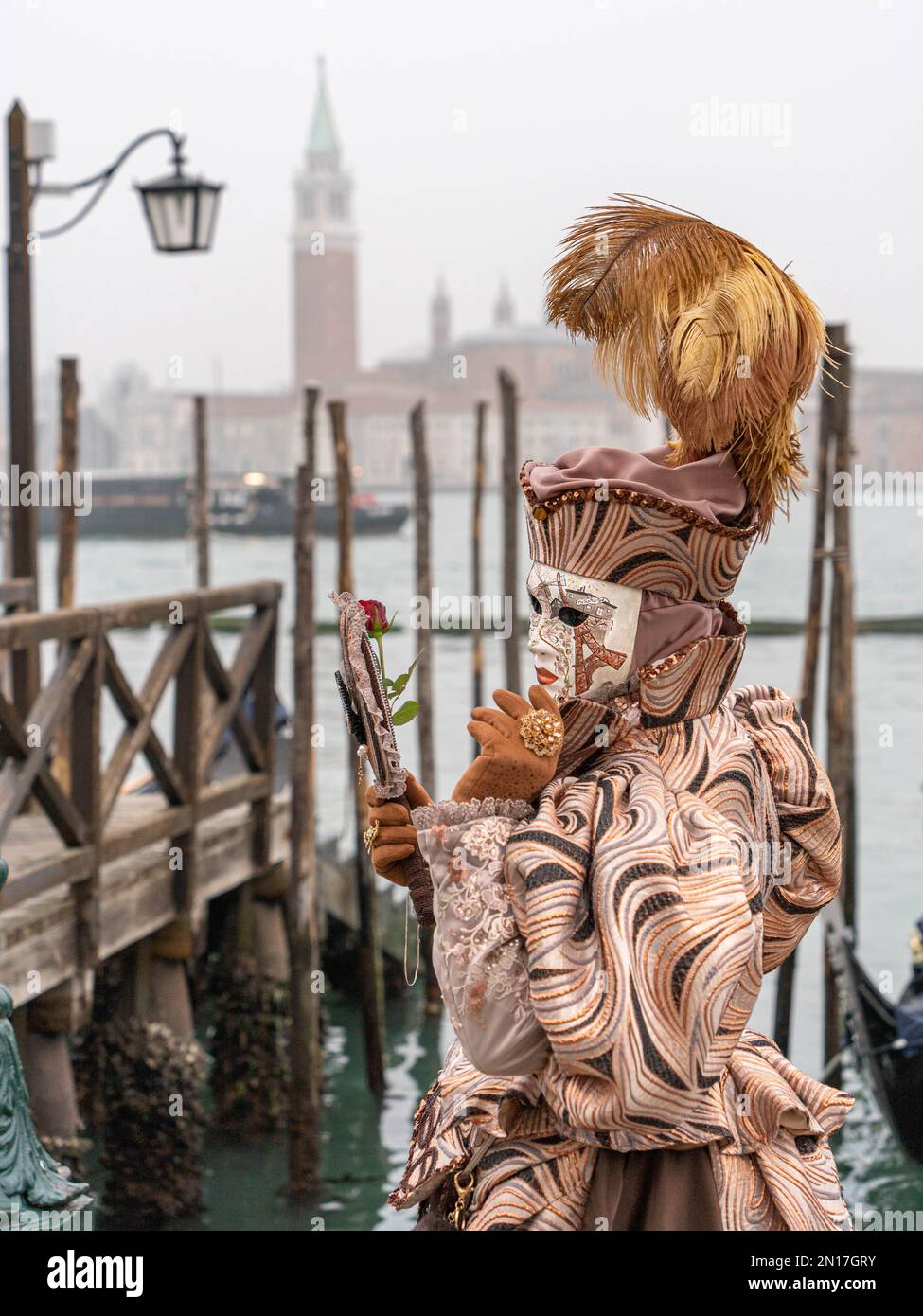 Una donna in costume da carnevale, una maschera, un cappello con piume, uno specchio e un fiore in mano, si trova sullo sfondo del molo e della Basilica Foto Stock