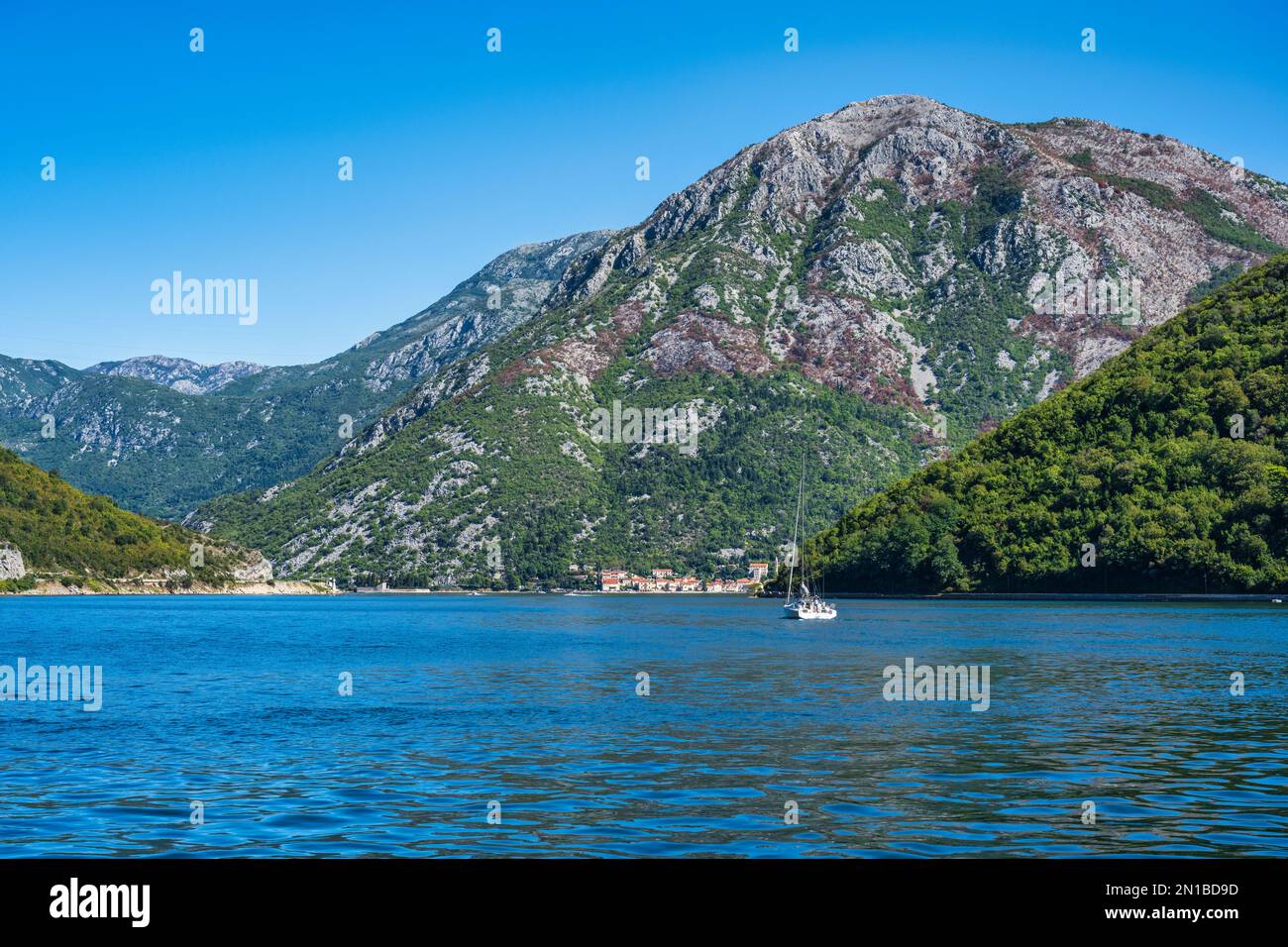 Lone yacht che naviga lungo lo stretto di Verige, la parte più stretta della baia di Cattaro, con vista in lontananza della città costiera di Perast in Montenegro Foto Stock