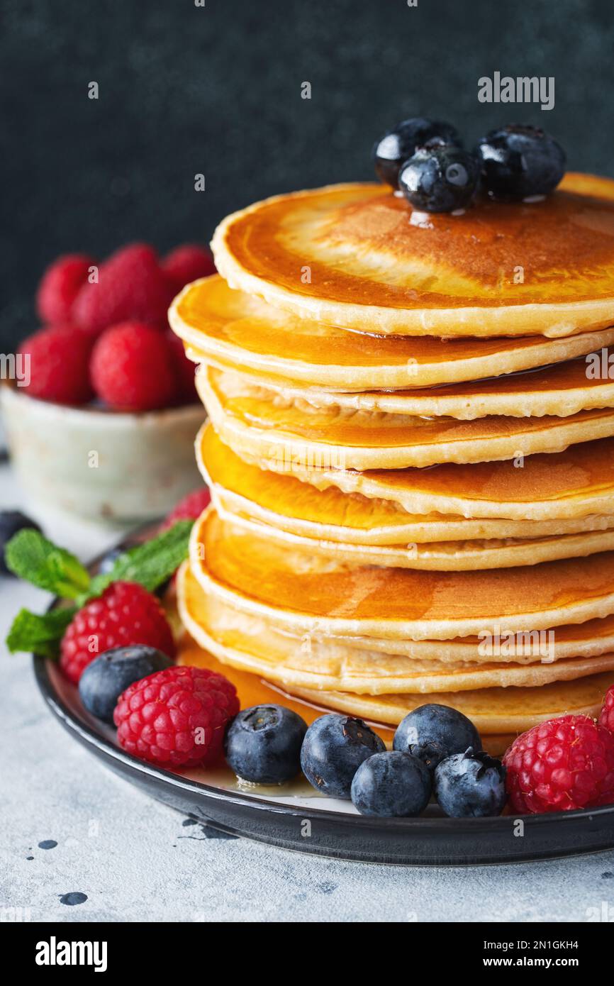 Primo piano deliziosi pancake, con mirtilli freschi, sciroppo di lampone e acero o miele su sfondo scuro. Lo sciroppo di acero dolce scorre da una pila di Foto Stock