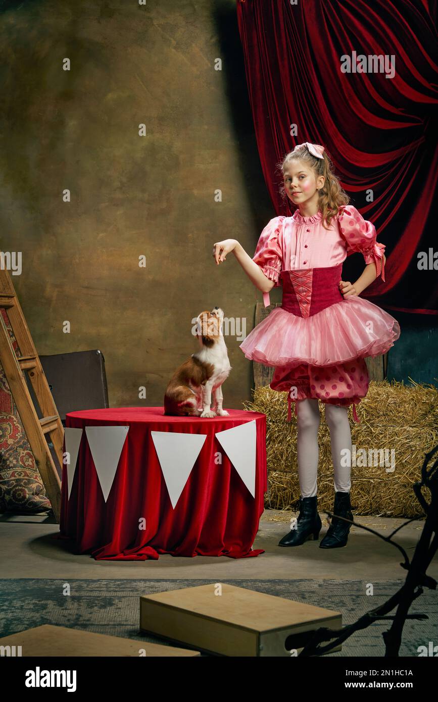 Fare prestazioni con il cane. Bella bambina in abito rosa festivo che si posa su sfondo scuro vintage circo. Vacanze, sogni, arte, moda Foto Stock