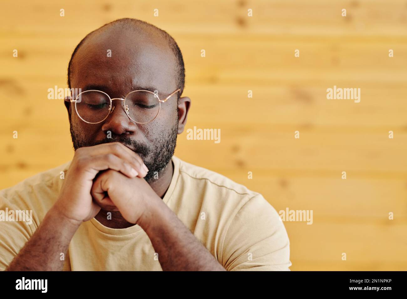 Giovane uomo nero in occhiali mantenendo gli occhi chiusi e mani aggrappate dalla bocca, concentrandosi sulla risoluzione di problemi gravi Foto Stock