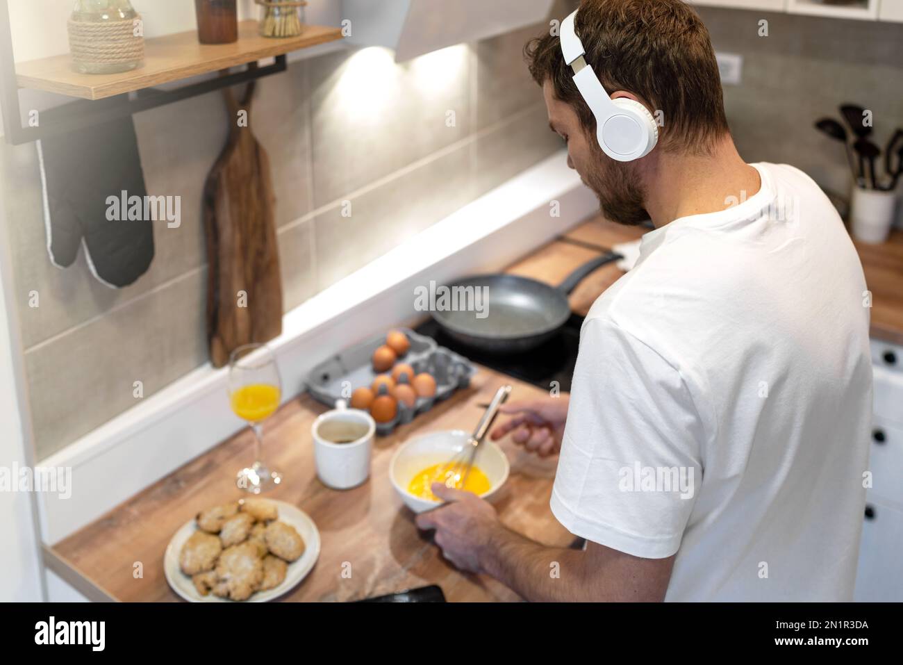 Uomo in maglietta bianca e cuffie in piedi in cucina frittata da cucina per la colazione e l'ascolto di musica. Foto Stock