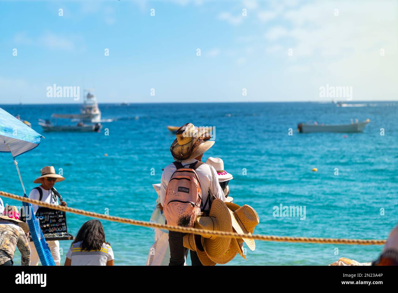 Un uomo messicano vende e indossa cappelli lungo la spiaggia pubblica di Playa Pública nella città turistica di Cabo San Lucas, Messico. Foto Stock