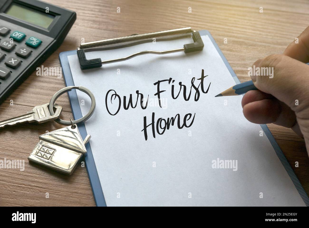 Messa a fuoco selettiva della calcolatrice, chiave della casa, una matita che tiene la mano che scrive una frase " la nostra prima casa" su una clipboard con carta su sfondo di legno. Foto Stock