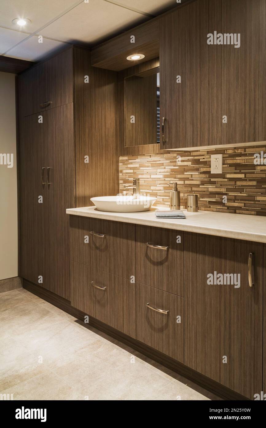 Armadietti in legno marrone e lavabo in stile ciotola bianca nella lavanderia, bagno nel seminterrato all'interno di una piccola casa. Foto Stock