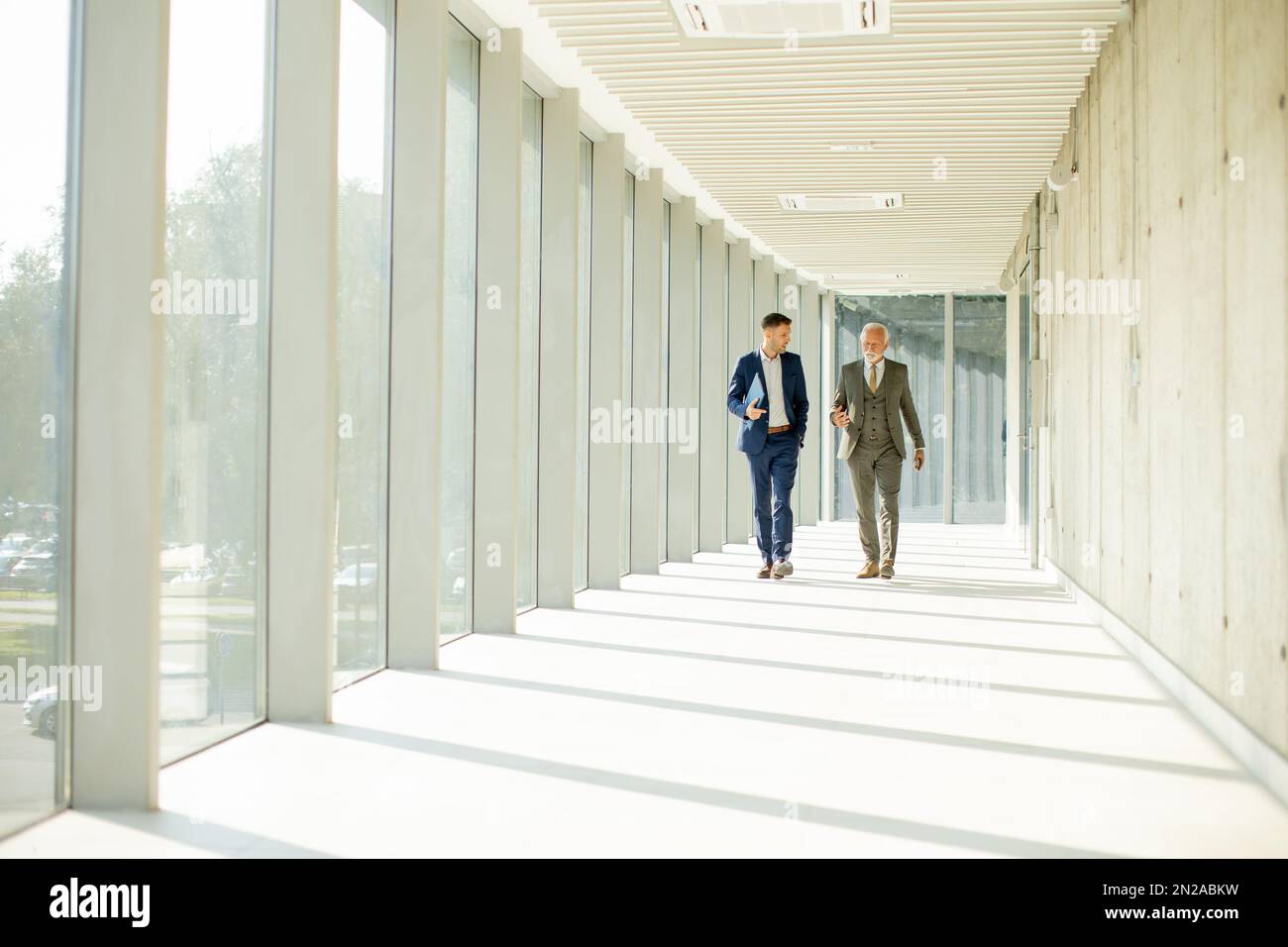 Un giovane e un uomo d'affari anziano camminano giù un corridoio dell'ufficio, profondo nella conversazione. Sono entrambi vestiti professionalmente, riflettendo il loro acum di affari Foto Stock