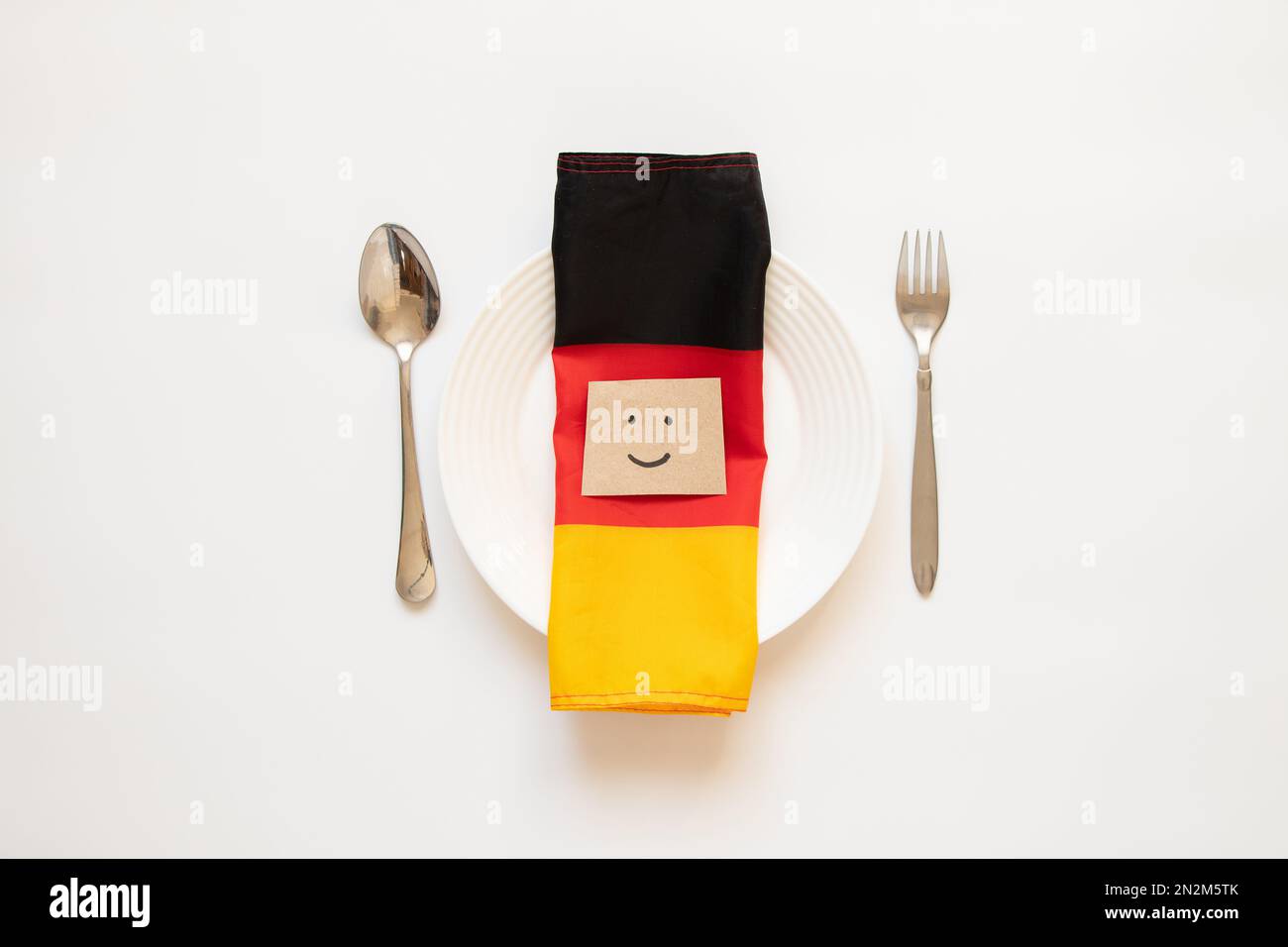La bandiera della Germania si trova su un piatto bianco accanto a un cucchiaio e una forchetta e una scheda con un sorriso dipinto su bianco, la bandiera nazionale Foto Stock