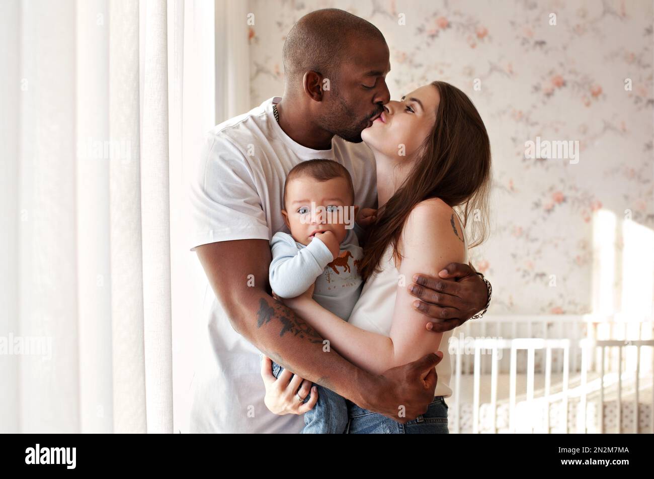 Felice famiglia multietnica che abbraccia e bacia a casa. Mamma e papà baciano mentre tengono il bambino tra le braccia, che guarda la fotocamera Foto Stock