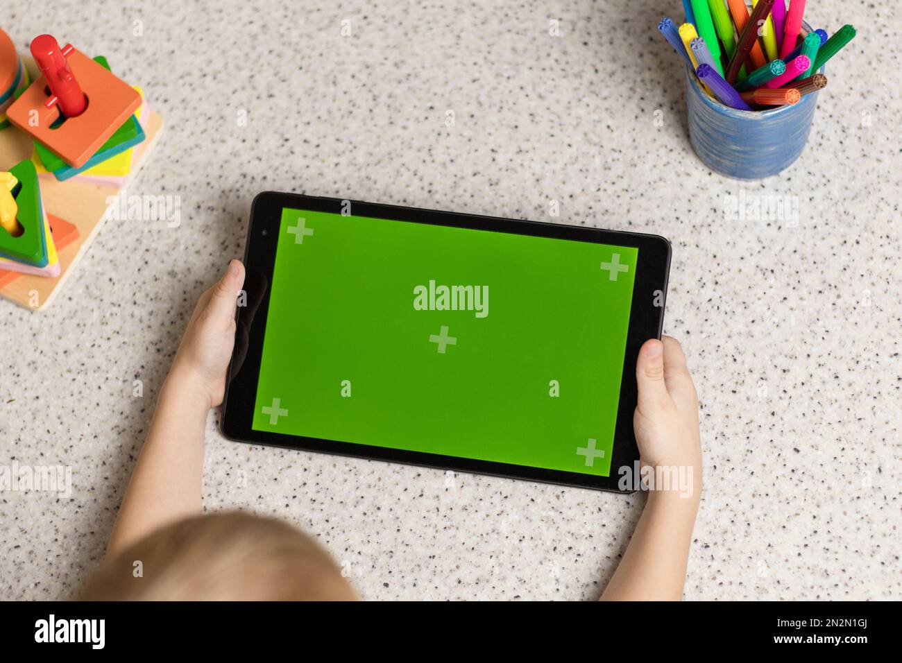 Le mani del bambino stanno tenendo un tablet con uno schermo verde. Chromakey. C'è un giocattolo e pennarelli colorati sul tavolo. Vista dall'alto. Foto Stock