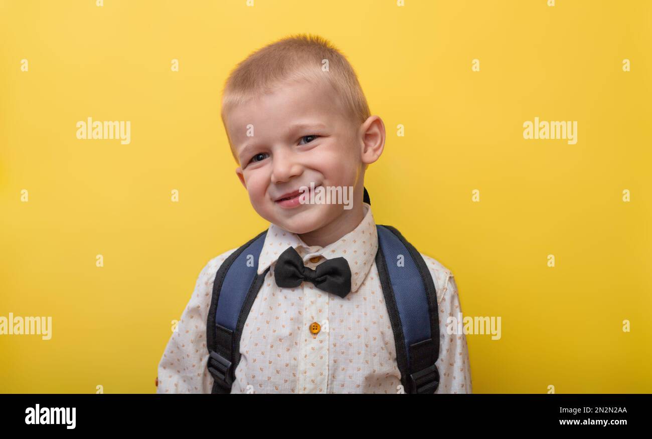 Ritorno a scuola. Un ragazzo biondo divertente con uno zaino in una camicia leggera su uno sfondo giallo sorride e guarda la macchina fotografica. Foto Stock