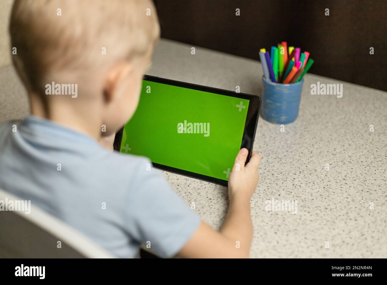 Il bambino è seduto al tavolo, gioca e guarda il tablet sullo schermo verde. Chromakey Foto Stock