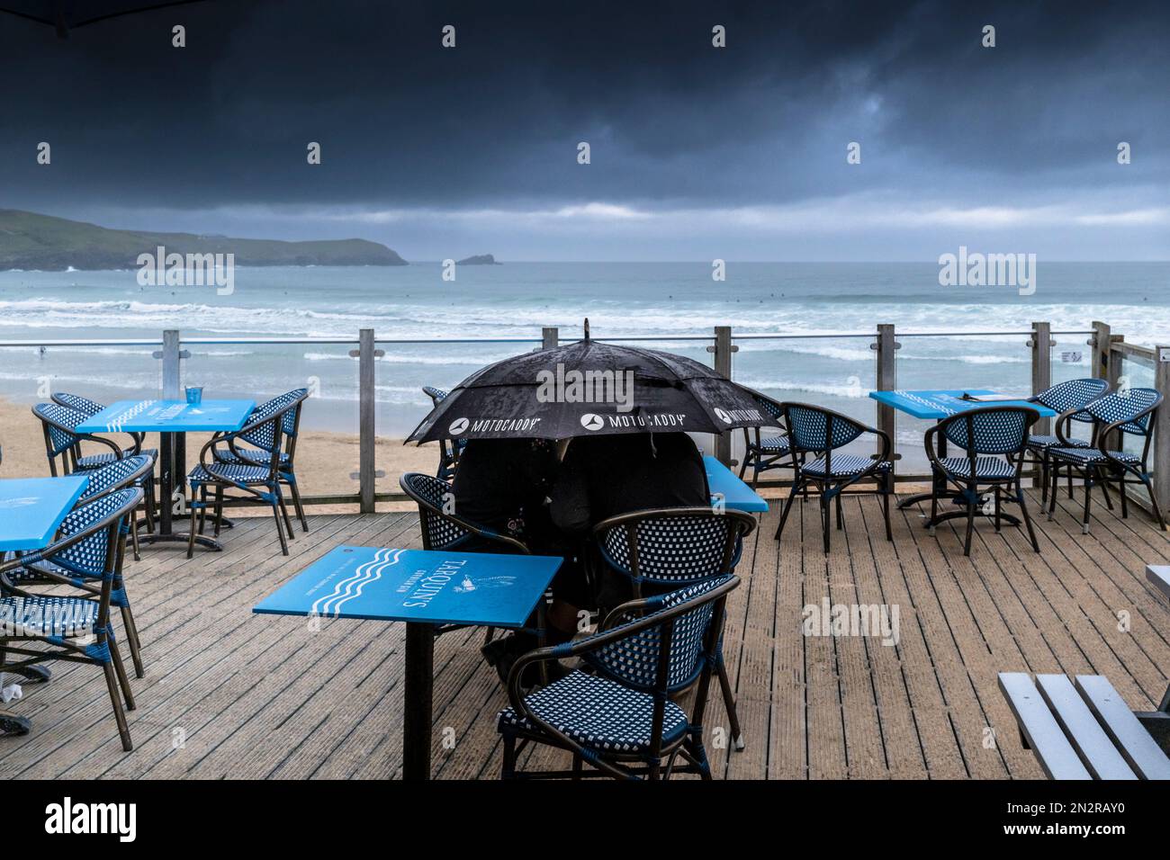 Meteo nel Regno Unito. Una coppia che si ripara sotto un ombrellone seduto ad un tavolo sulla terrazza esterna del Fistral Beach Bar che si affaccia sulla spiaggia di Fistral Foto Stock