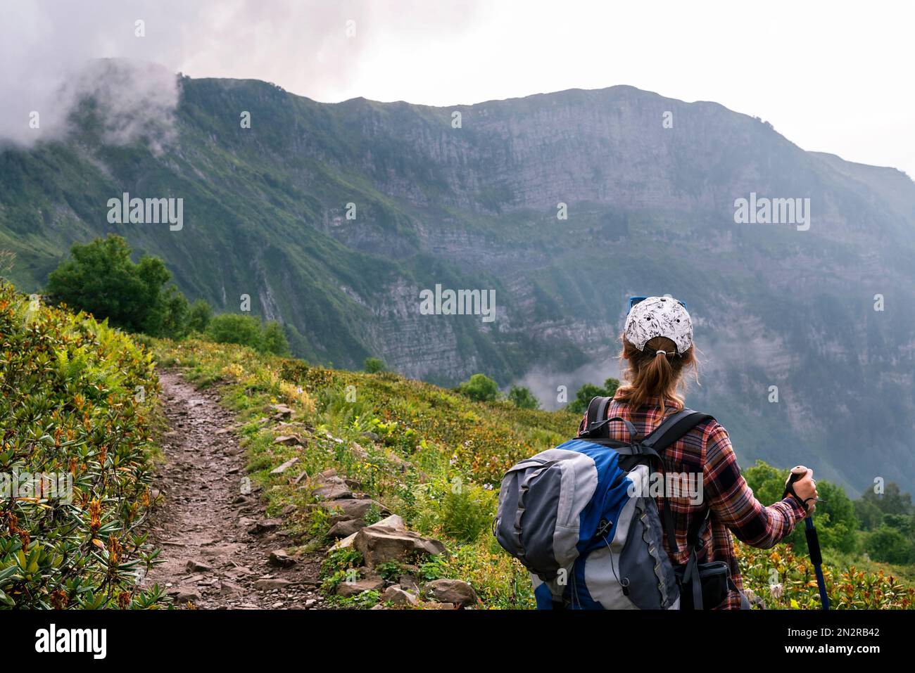 vista posteriore della giovane donna in camicia a quadri e berretto con grande zaino con bastoni da trekking camminando lungo il sentiero di montagna in estate escursioni sano lif attivo Foto Stock