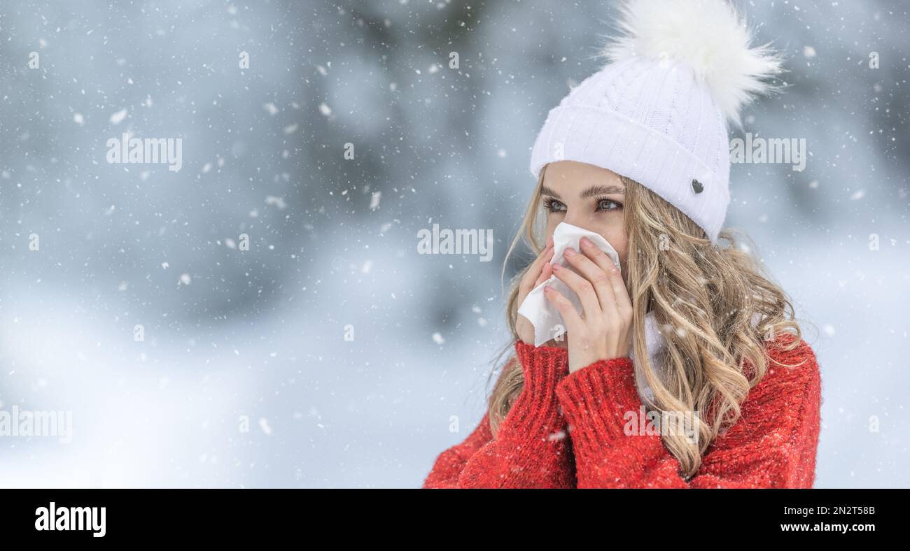 Giovane donna con sintomo allergico naso che soffia. Una bambina malata in un caldo maglione a maglia e una cuffia di lana con un fazzoletto in un parco invernale. Foto Stock